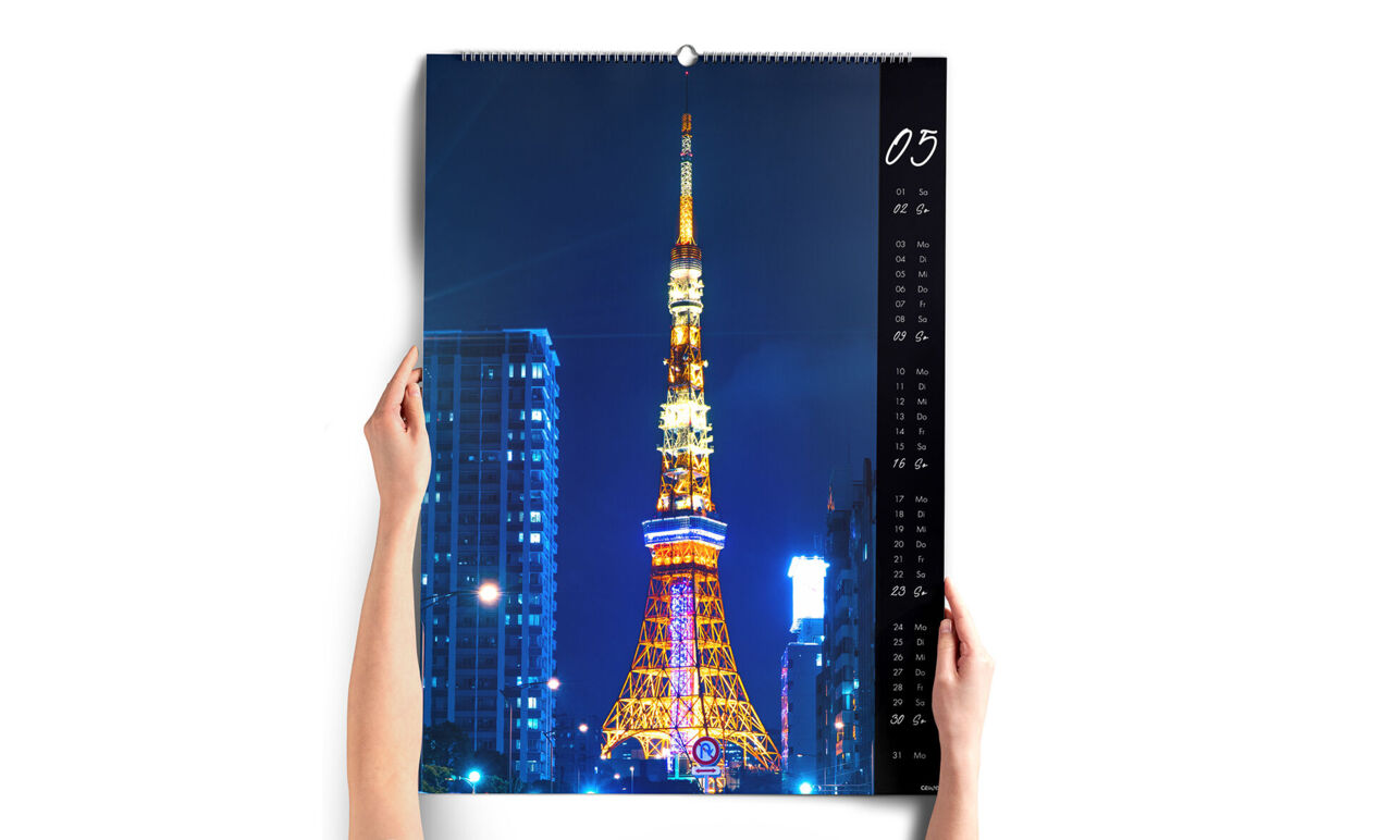Zwei Hände halten den XXL-Wandkalender im Hochformat vor sich. Das Kalenderblatt zeigt einen beleuchteten Turm in einer Großstadt bei Nacht
