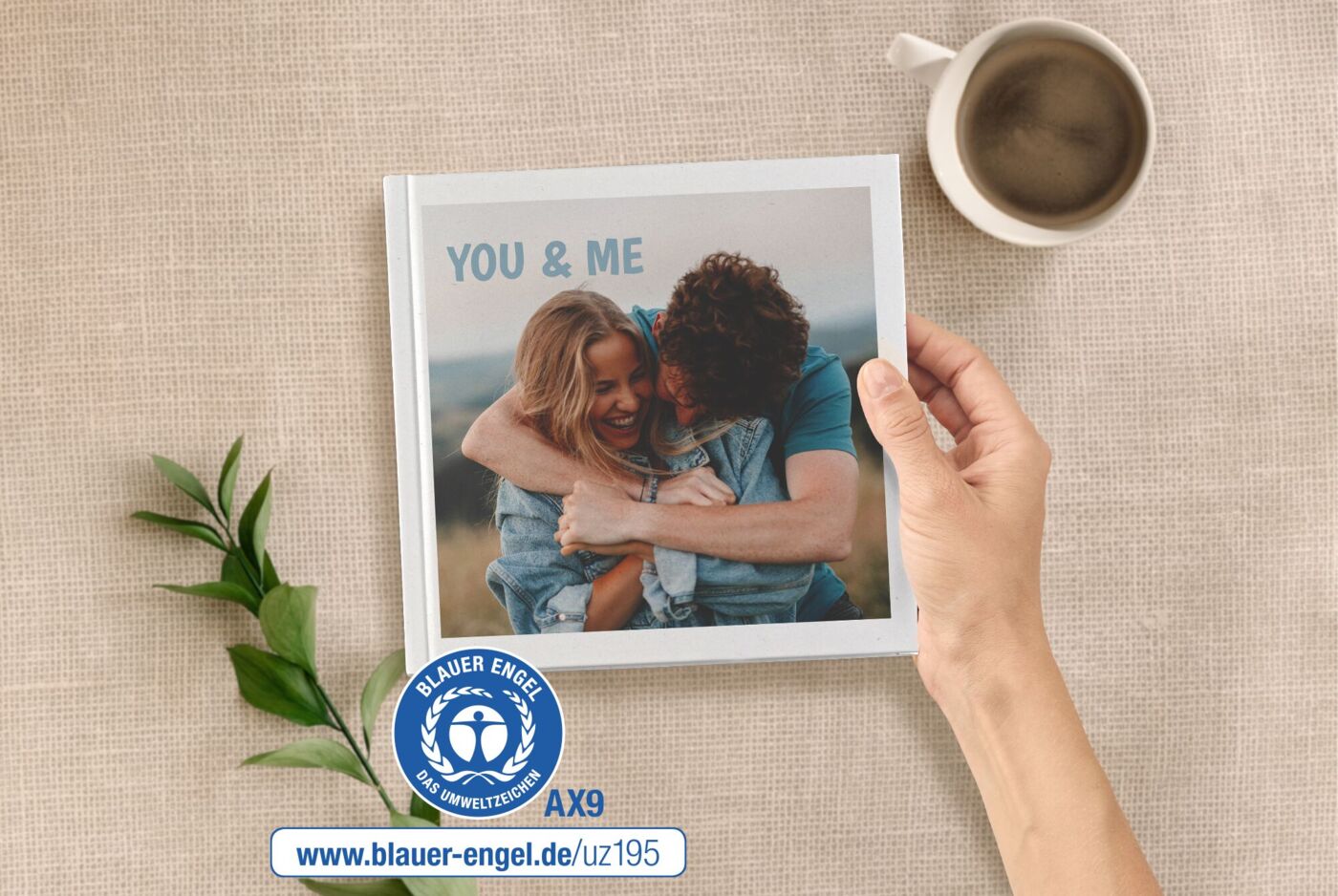 Ein Fotobuch im Format 20x20 wird von einer Hand gehalten. Auf dem Cover ist ein Paar zu sehen. Daneben ist der Blaue Engel.