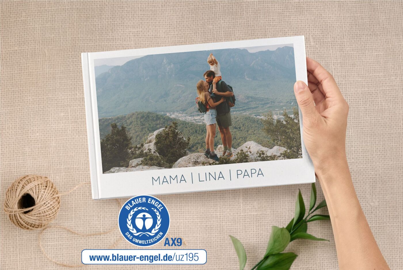 Eine Hand hält ein A4 Quer Fotobuch. Auf dem Cover ist eine Familie beim Wandern abgebildet. Daneben ist der Blaue Engel.