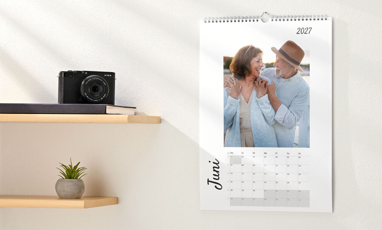 Kalender im Hochformat mit Spiralbindung, weißem Design und Foto eines älteren Paares, hängt an der Wand neben einem Regal.
