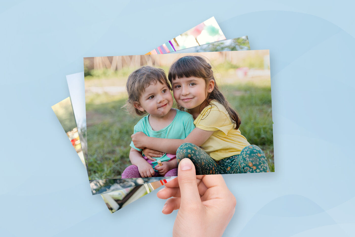 Eine Hand hält einen Stapel Fotoabzüge. Auf dem vordersten Foto sind zwei Kinder auf einer Wiese abgebildet, die sich umarmen