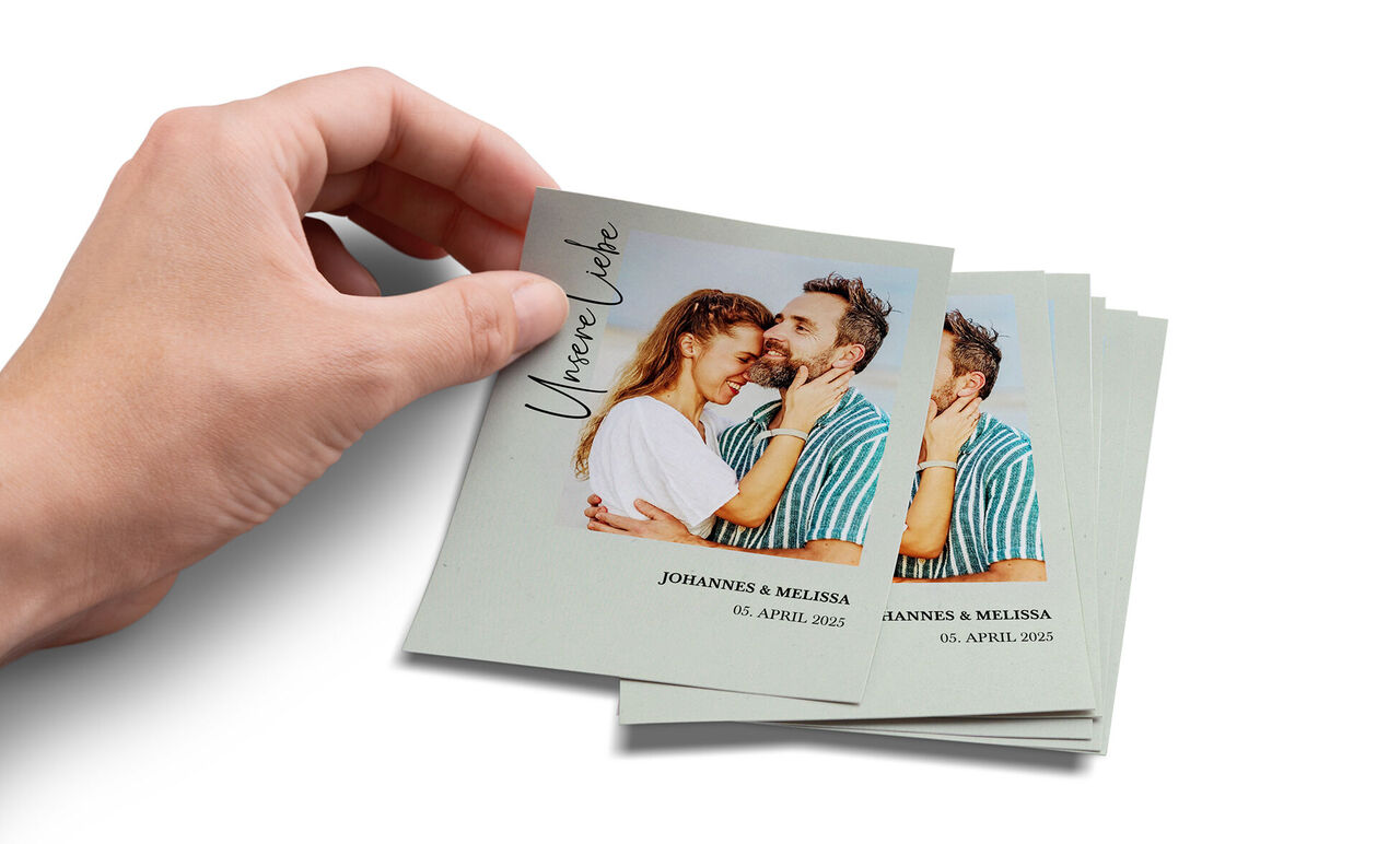 Eine Person hält ein personalisiertes Foto mit dem Aufdruck "Unsere Liebe" in der Hand. Das Foto zeigt ein glückliches Paar, das sich umarmt, und die Schriftzüge beinhalten die Namen und das Datum.