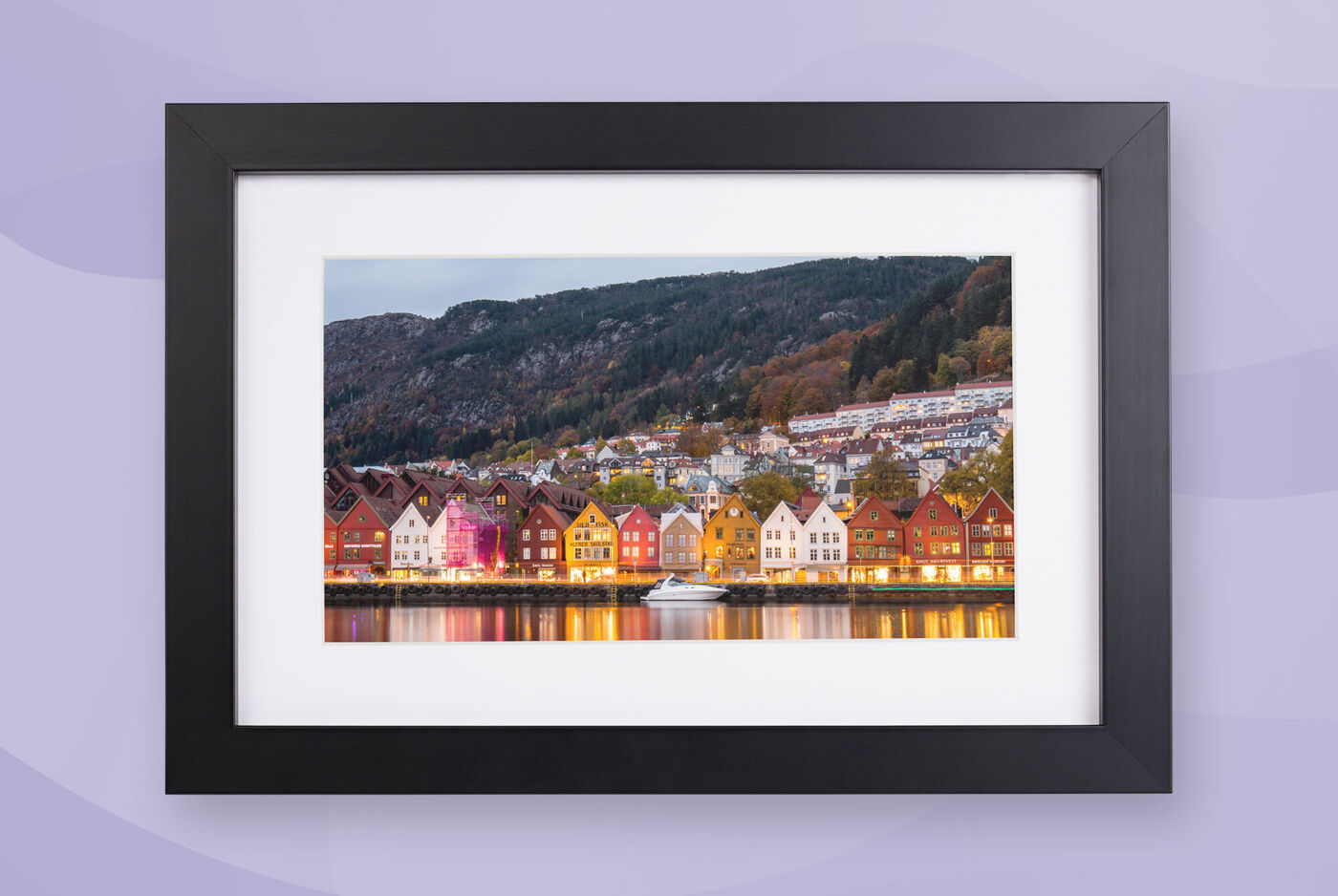 Gerahmtes Foto einer norwegischen Hafenstadt bei Dämmerung: Bunte Häuser leuchten am Wasser, dahinter ein Berg. Lichter spiegeln sich im Haf