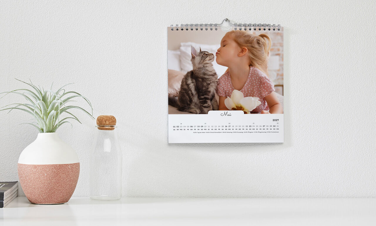 Quadratischer Kalender mit weißem Design, mit Foto von Mädchen mit Kätzchen, hängt an der Wand über einer Anrichte.