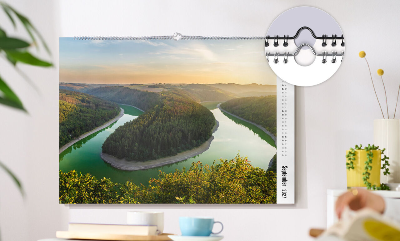 XXL-Wandkalender im Format 50 x 70 cm hängt an einer Wand. Das Kalenderblatt zeigt einen Fluss, der durch eine Berglandschaft verläuft