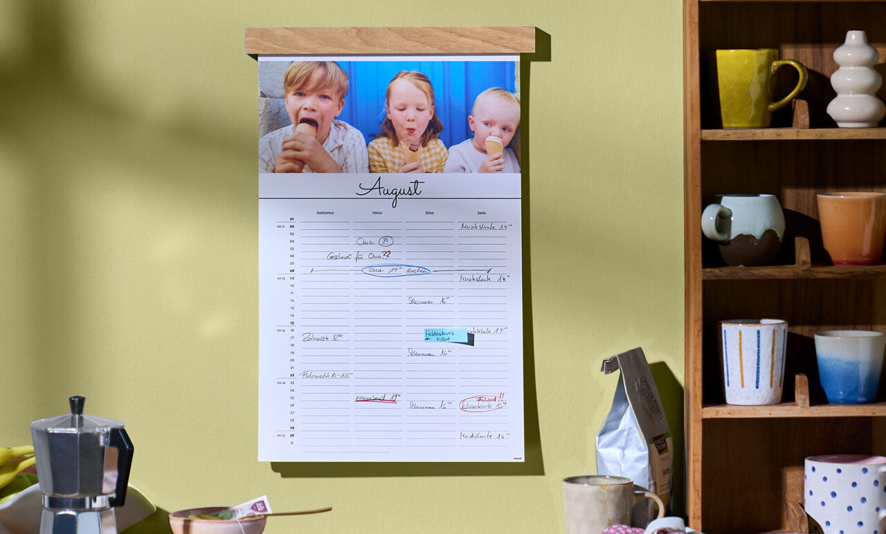 An einer Wand hängt ein Kalender mit Holzleiste neben einem Regal. Das Kalenderblatt zeigt ein Kinderfoto und mehrere Spalten für Eintäge