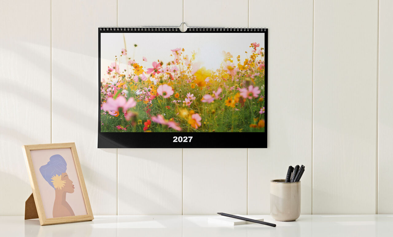 Kalender im Querformat mit Spiralbindung und einfachem, schwarzem Design, mit Foto einer Blumenwiese, hängt über Sideboard.