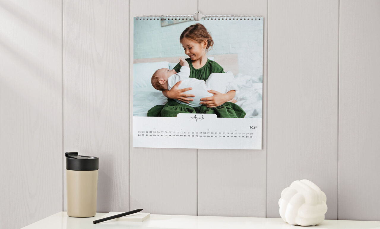 Quadratischer Kalender mit weißem Design und Foto von einem Mädchen mit Baby im Arm, hängt an der Wand über einer Anrichte.