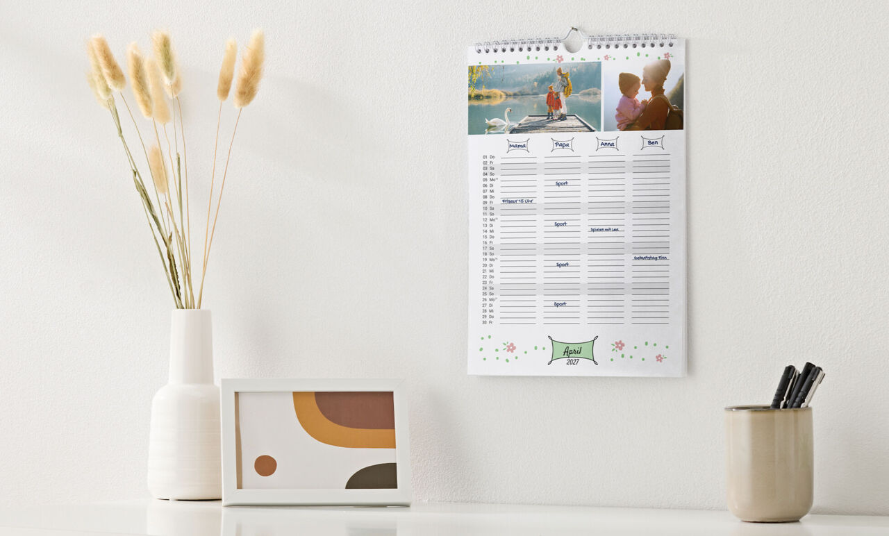 Familienkalender mit Spiralbindung, Kinderfotos und einem Design mit Spalten für vier Personen, hängt über einer Anrichte.