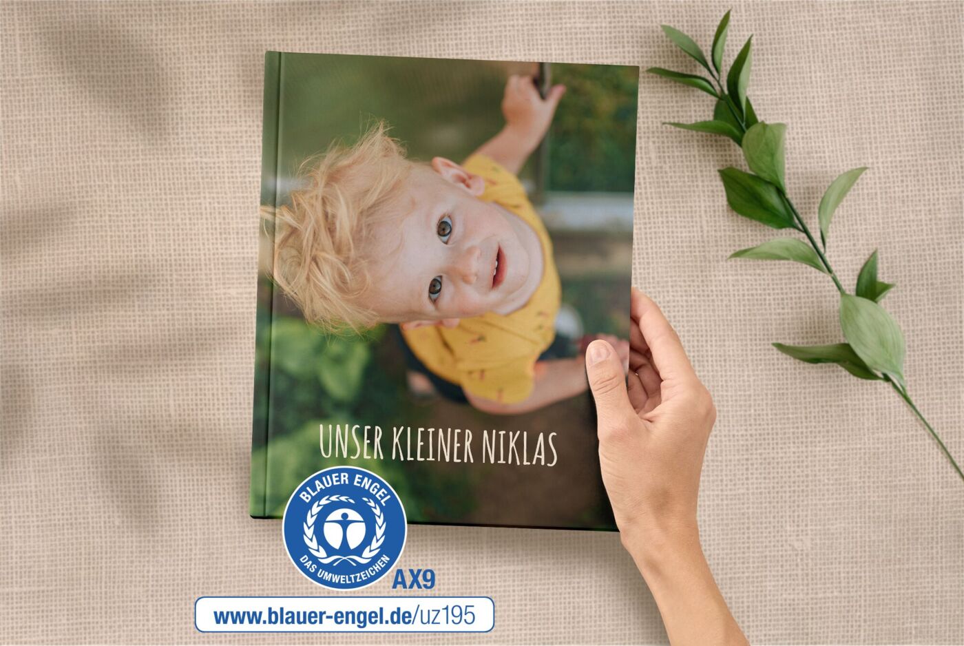 Eine Hand hält ein A4 Hoch Fotobuch. Auf dem Cover ist ein kleiner Junge abgebildet. Daneben ist der Blaue Engel.