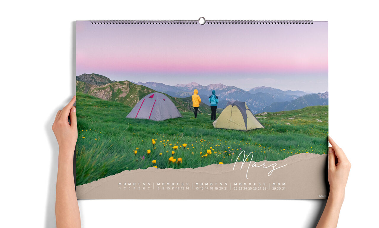 Zwei Hände halten den XXL-Wandkalender im Querformat. Das Blatt zeigt zwei Personen in bunten Jacken neben Zelten auf einer Bergwiese.
