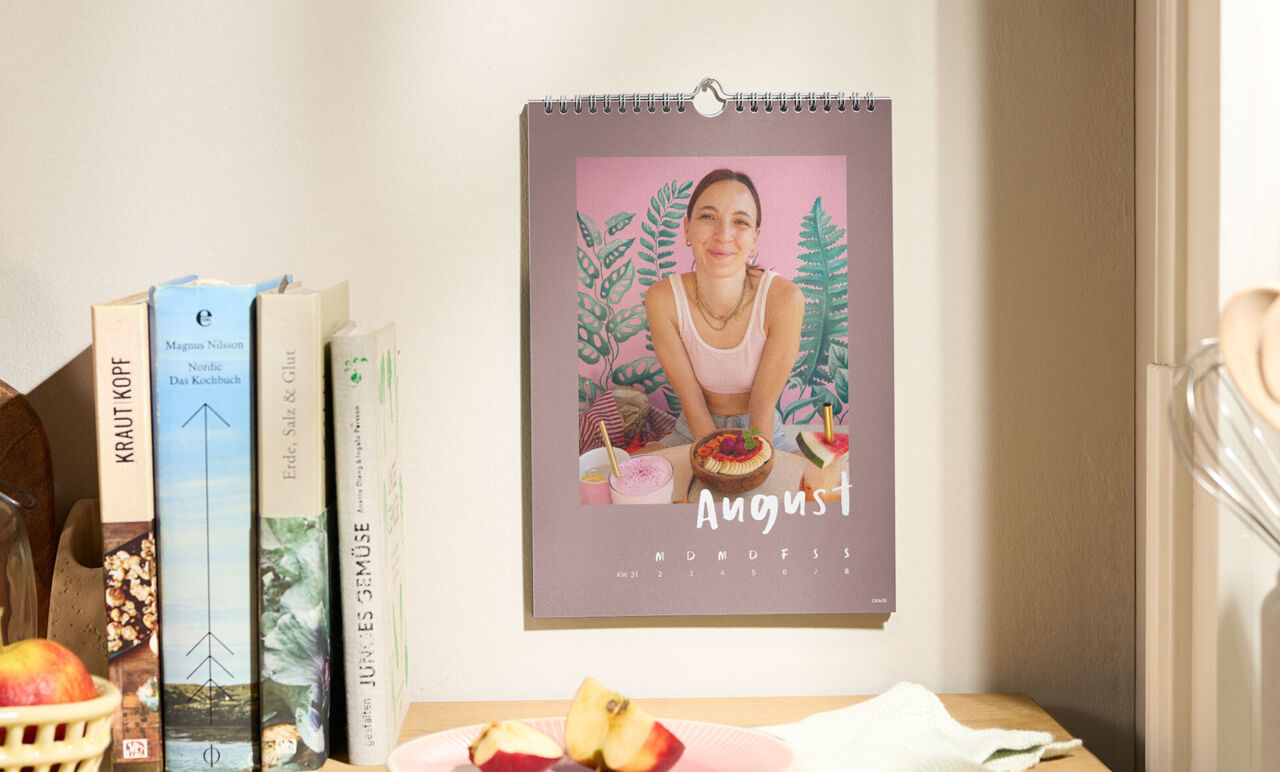 Ein Wandkalender im A4-Format hängt über einem Bücherregal. Das Augustblatt zeigt ein Foto eines Mädchens und eine Wochenansicht