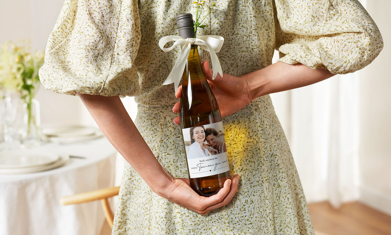 Eine Person hält eine Flasche Wein mit einem dekorativen Etikett und einer Schleife.